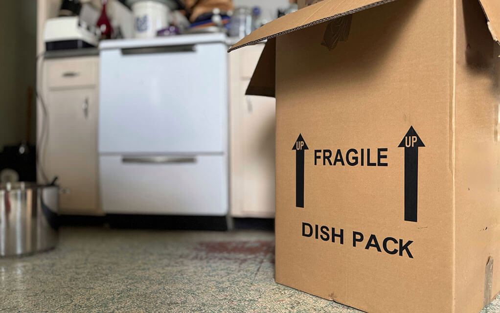 A box with the words "FRAGILE" and "DISH PACK" sits in a kitchen waiting to be packed as the kitchen oven sits in the background