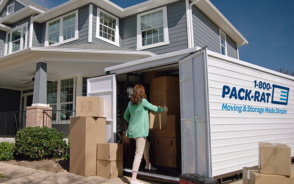 A woman is packing boxes into her 1-800-PACK-RAT portable storage container sitting in her driveway as boxes are stacked nearby the container on a bright, clear day.