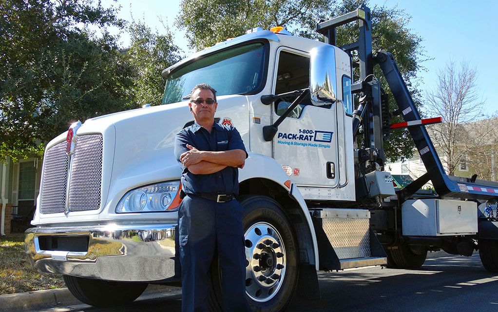 An older but confident looking male 1-800-PACK-RAT driver stands with his arms crossed in front of his unloaded mini-mover truck